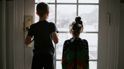 Back view of two young children looking through window of home