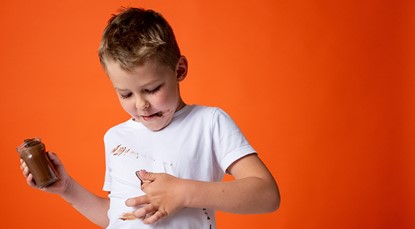 Young boy wearing a white t-shirt, smearing chocolate spread on his shirt while holding a jar of it in the other hand.