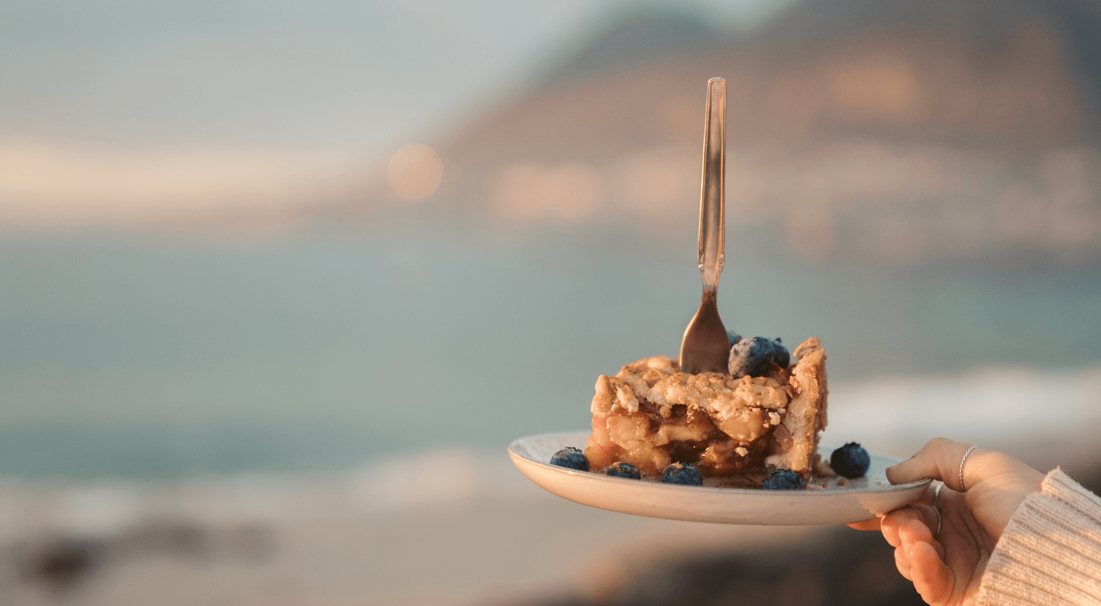 Person holding a slice of cake on a plate against a coastal backdrop.