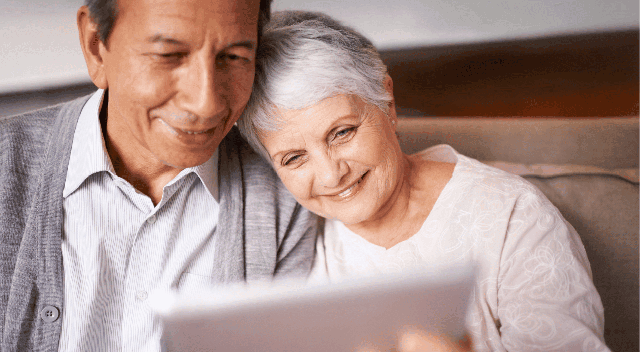 Elderly couple sitting on a couch together, smiling and looking at a tablet