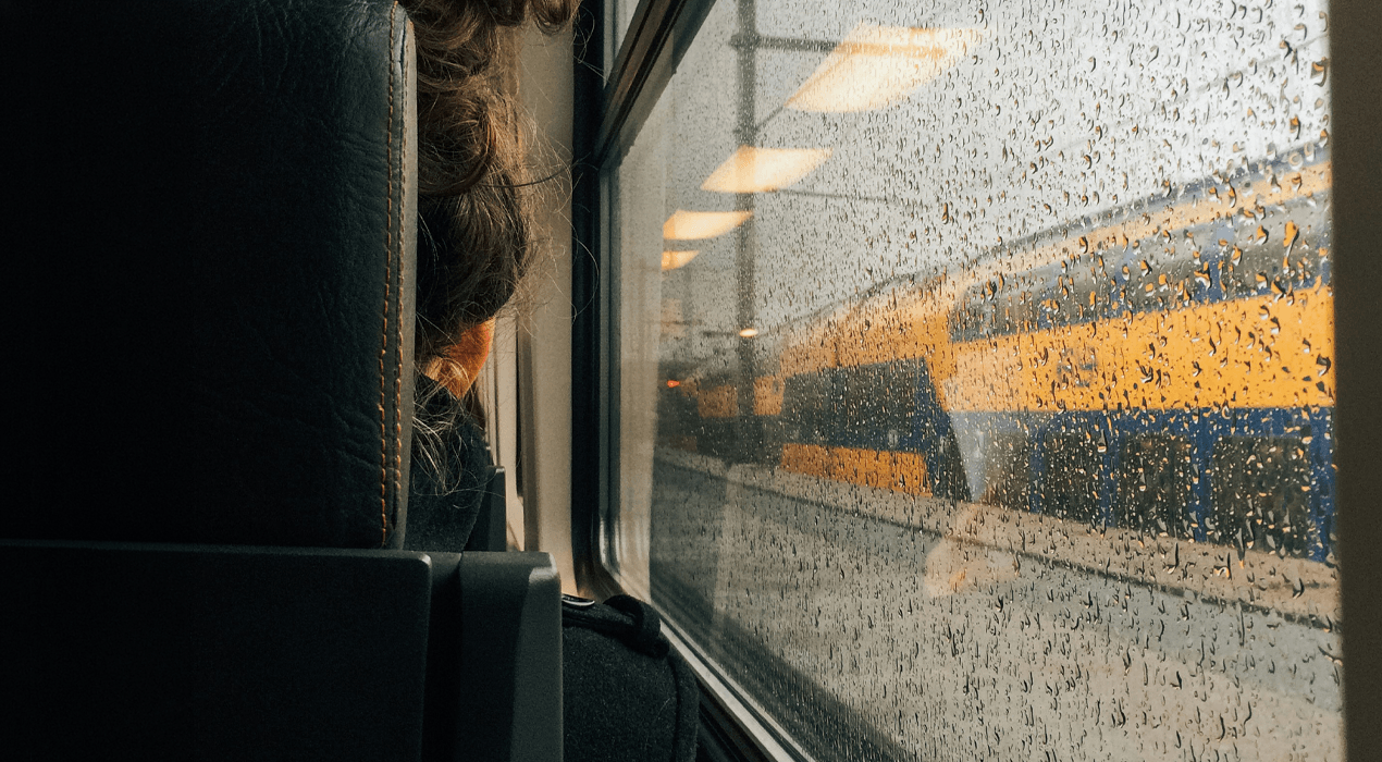 Rear view of a woman sitting looking out a train window, with the window covered in rain drops
