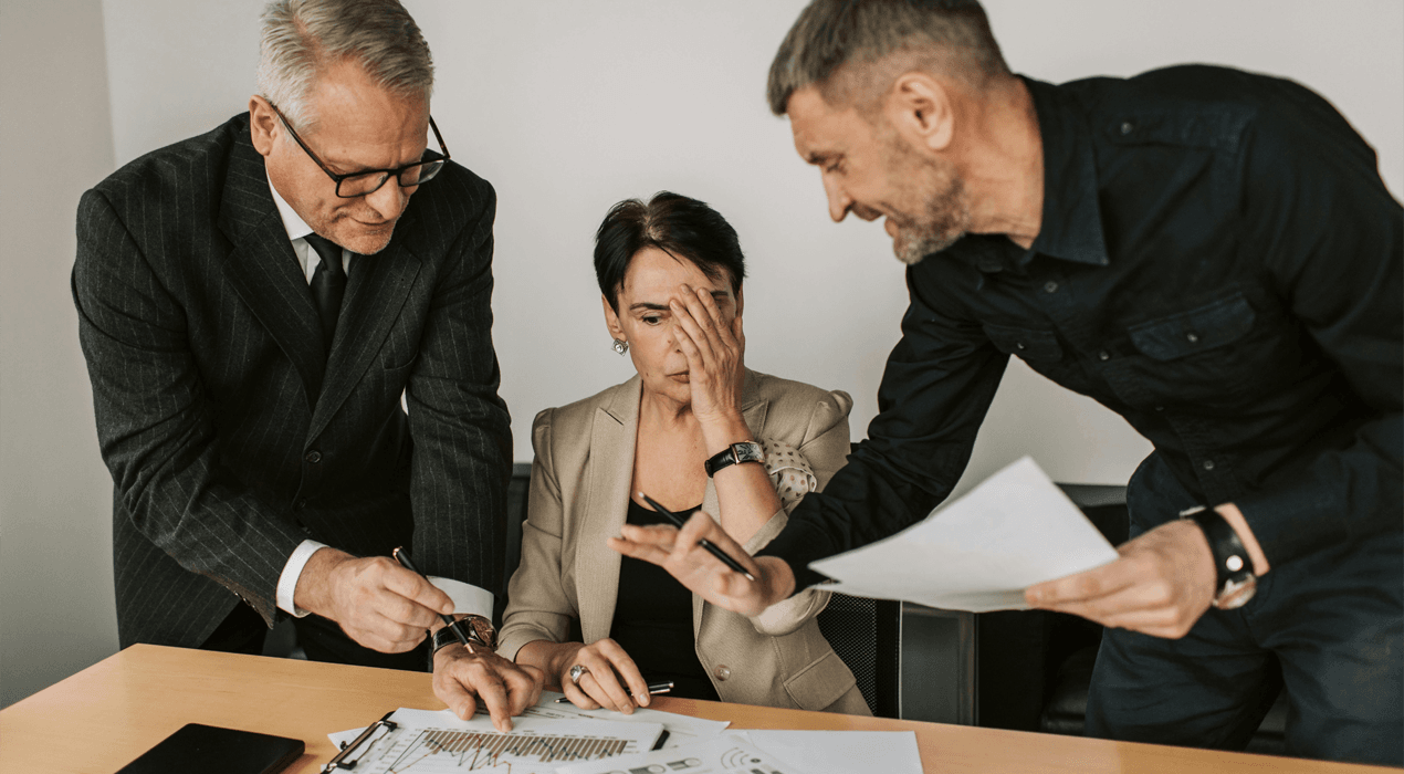 Woman sitting at a desk looking overwhelmed, two men stand either side pointing to papers / charts in front of her