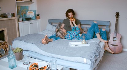 Young couple in their home, lying on bed eating pizza