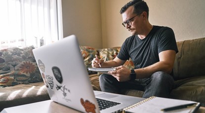 Man in black T-shirt and jeans sitting on couch, holding pad of paper in front of laptop