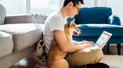 Guy wearing white tshirt and chinos leaning against couch on the floor, with dog using laptop