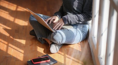 Woman in grey jumper and jeans sitting on floor of home using laptop