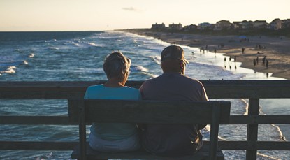 Back of middle aged couple sitting on bench looking at beach