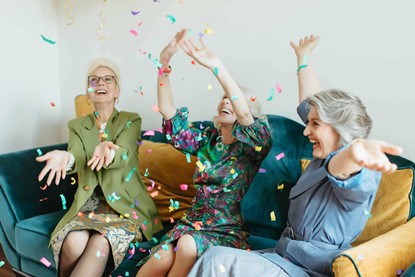 Three older women sitting on a couch, celebrating and throwing confetti in the air