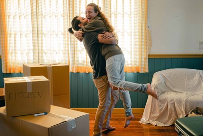 Young couple smiling and hugging in the middle of a lounge, between a pile of moving boxes and a couch