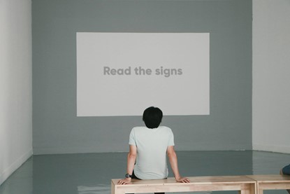 Man sitting on a wooden bench inside a gallery looking up at a wall with the words printed "Read the signs".