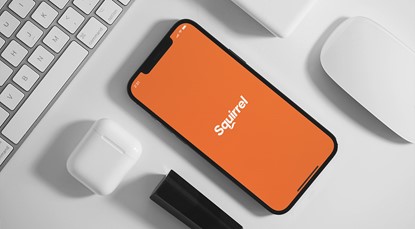 iPhone laying on flat surface, displaying the loading page for the Squirrel Money app (orange background with white Squirrel logo in the middle). Phone surrounded by other tech gadgets, including a keypad, mouse and Airpods case.