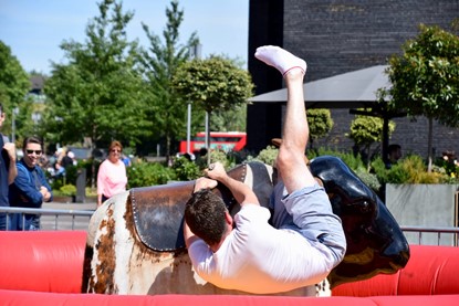 Man falling off a mechanical bull, as several onlookers watch on. Man wears white t-shirt and light blue shorts, with white socks.