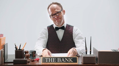 Smug looking man sitting at a desk, behind a nameplate that says "The Bank". The man is dressed in a white collared shirt, black bowtie and a dark maroon waistcoat.