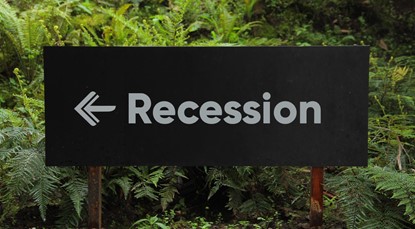 Black sign with a white arrow pointing left, and the word "Recession", against a plant backdrop.